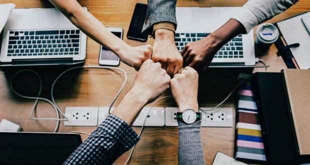Teamwork and unity concept with diverse hands joining in a fist bump over a desk with laptops and office supplies.
